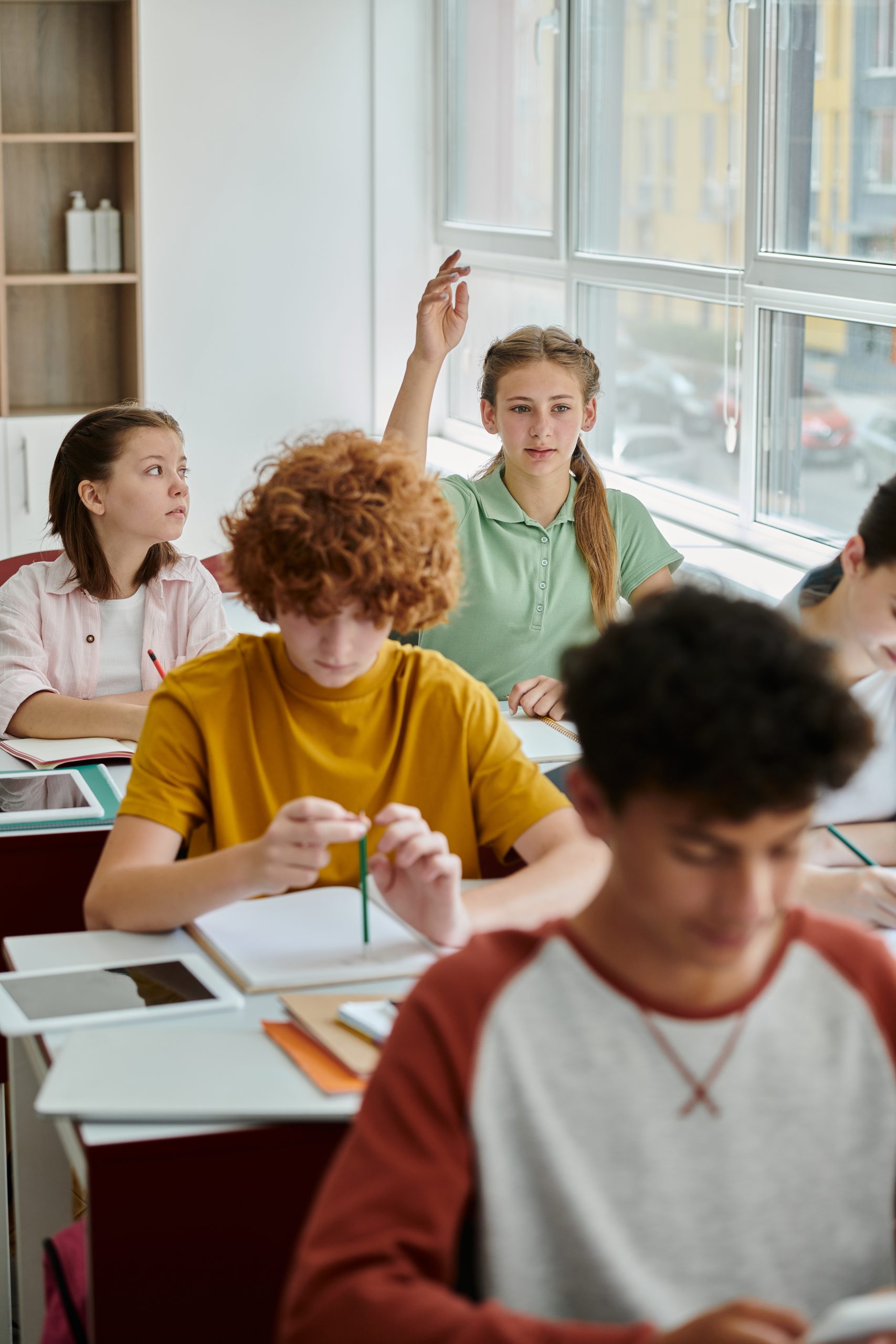 Teenage schoolgirl raising hand and talking near devices and classmates during lesson in school